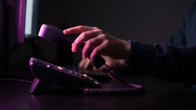 A hand lifting a desk phone handset in a dark office, showing an AI receptionist handing a call to a person