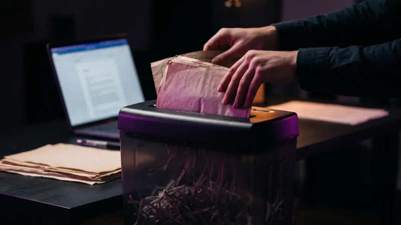Hands feeding old paper records into a shredder beside a laptop, a small business scaling down data it no longer needs
